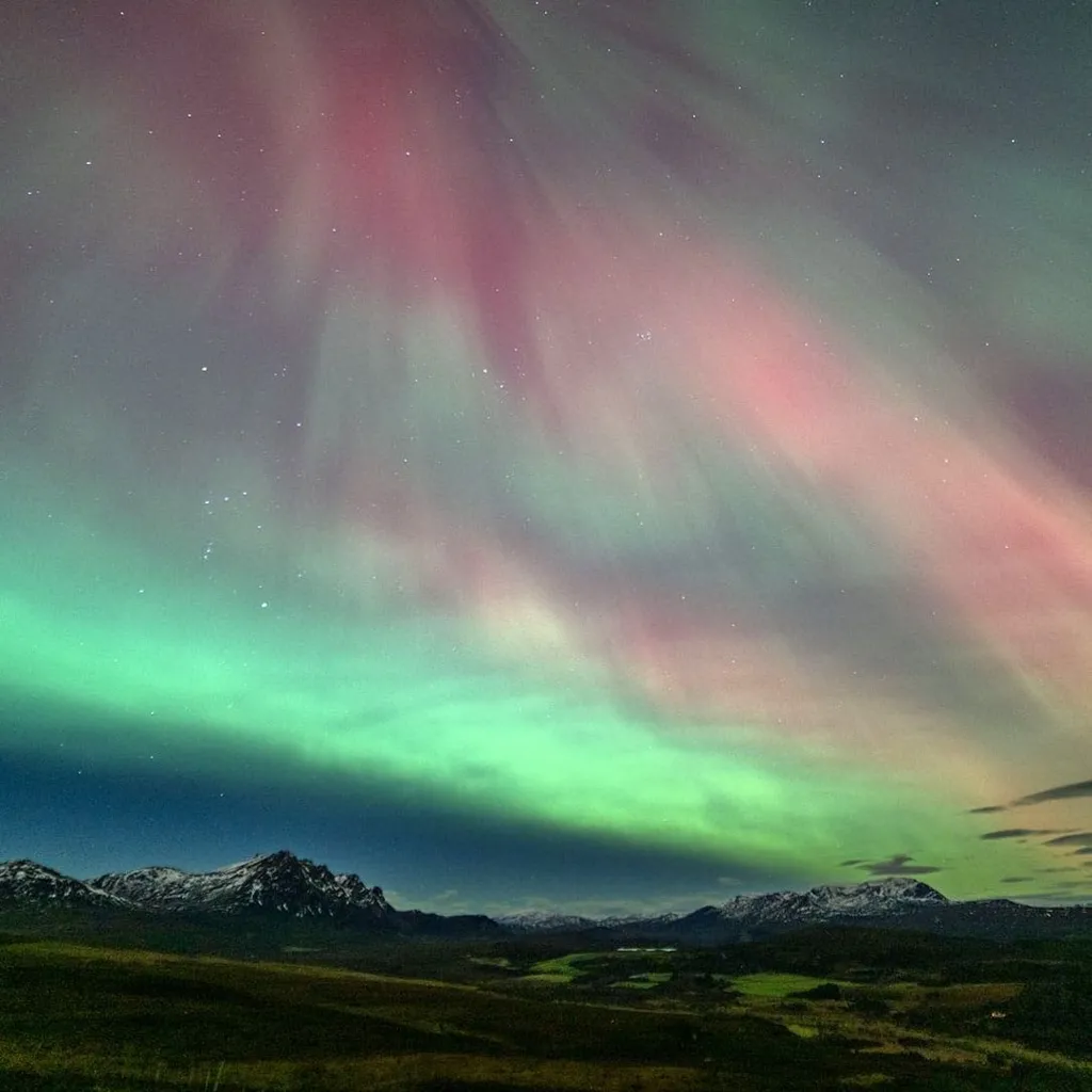 The Northern Lights in the north coast of Scotland. There are green and red stripes across the sky with snow-capped mountains below. 