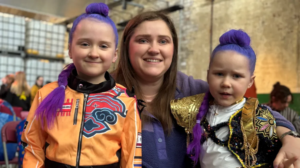 Two children are dressed as Kpop Demon Hunters. They have purple hair and and are wearing colourful costumes. They are standing at either side of their mum who has dark hair and is wearing a purple jumper.