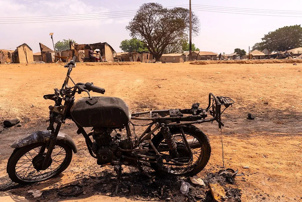 The charred frame of a burnt motorbike sits on dry, yellow grass near a small rural village.