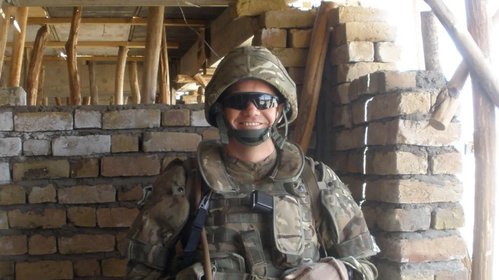 Wearing combat uniform and dark glasses, Lt Col Teeton smiles at the camera from the interior of a partly-constructed building in Afghanistan
