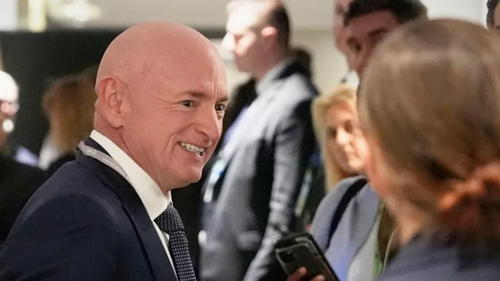 Senator Mark Kelly, in a dark blue suit and patterned tie, smiles and turns toward the camera in front of a group of people