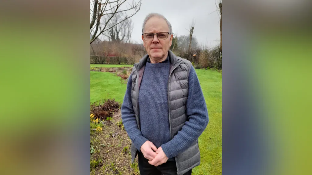 Wilson Beggs is standing outside with grass and flowers behind him. He's wearing a navy vest and jumper. He wears glasses. 