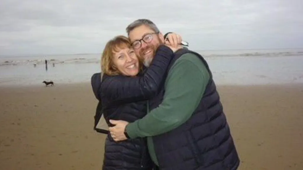 Carolyn, wearing a black puffer coat, hugs her husband - who wears a navy puffer and green jumper - on a windswept beach