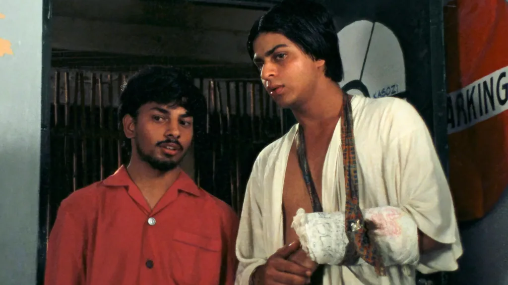 A young Shah Rukh Khan (on the right), seen in a white robe and a plaster and a sling in his right hand which seems to be broken. He is standing next to a shorter man, wearing a red t-shirt.