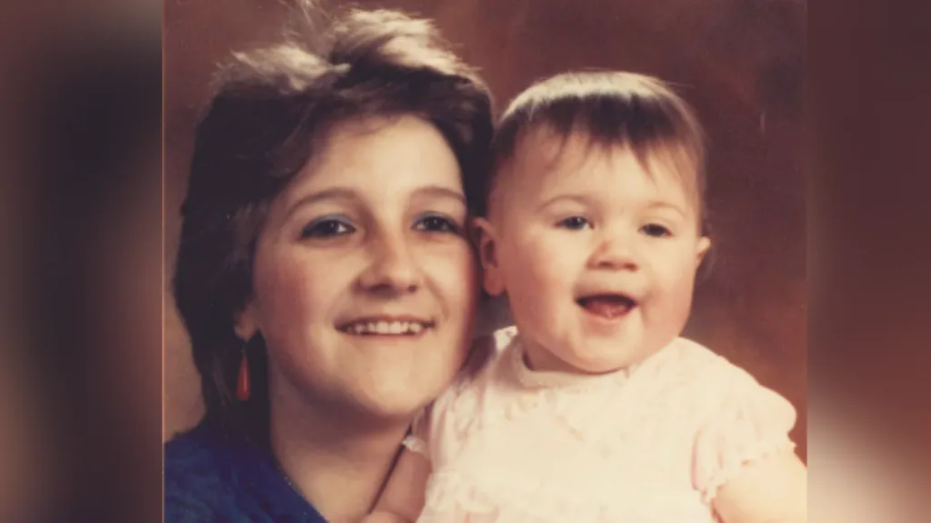 Pauline Watkins as a young mother. She has Short light brown hair and is wearing makeup and drop red earrings. She wears a dark blue top. She holds baby Jemma who is less than a year old and is wearing a white or pink ruffly top. The picture is from a professional studio. Both look to the right of the picture.