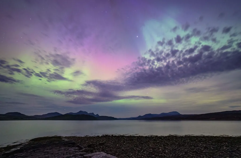 The Northern Lights in the north coast of Scotland. There are green and purple stripes across the sky with mountains and the sea below. 