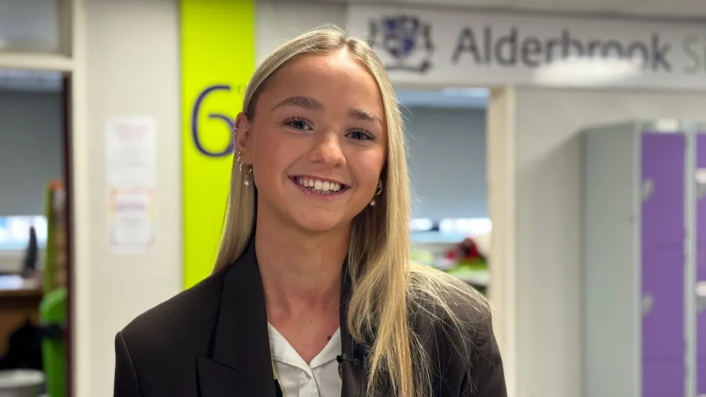 Year 13 student Jasmine has blonde hair and brown eyes. She wears a black blazer and white shirt and is smiling. She is standing in the sixth form corridor with some of the common rooms behind her.