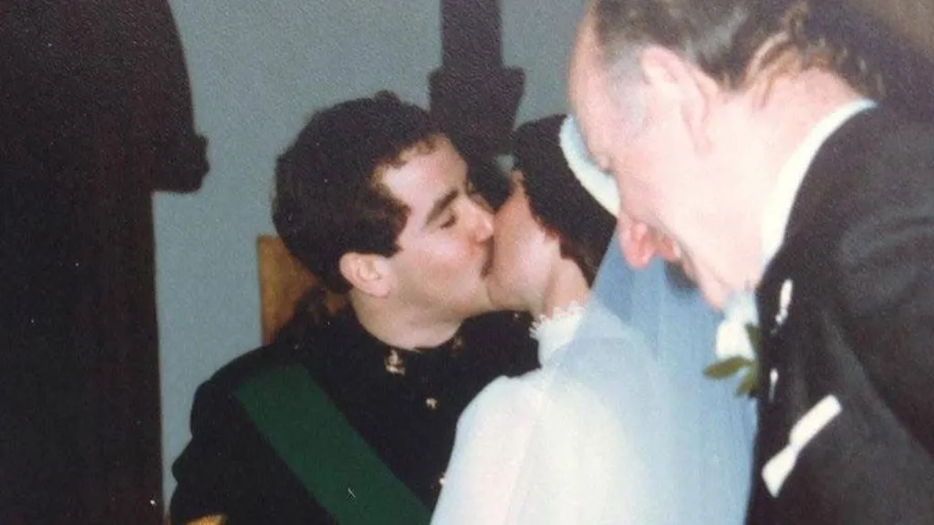 There are four people in this photograph. At its centre is a bride and groom kissing on their wedding day. The groom is wearing a black military uniform. The bride - who has her back to the camera - has a high necked white dress and a white veil pinned to a headdress. They each have dark hair. To the left of the groom is the Cannon, who married them. To the right of the bride is a wedding guest in a black suit. 