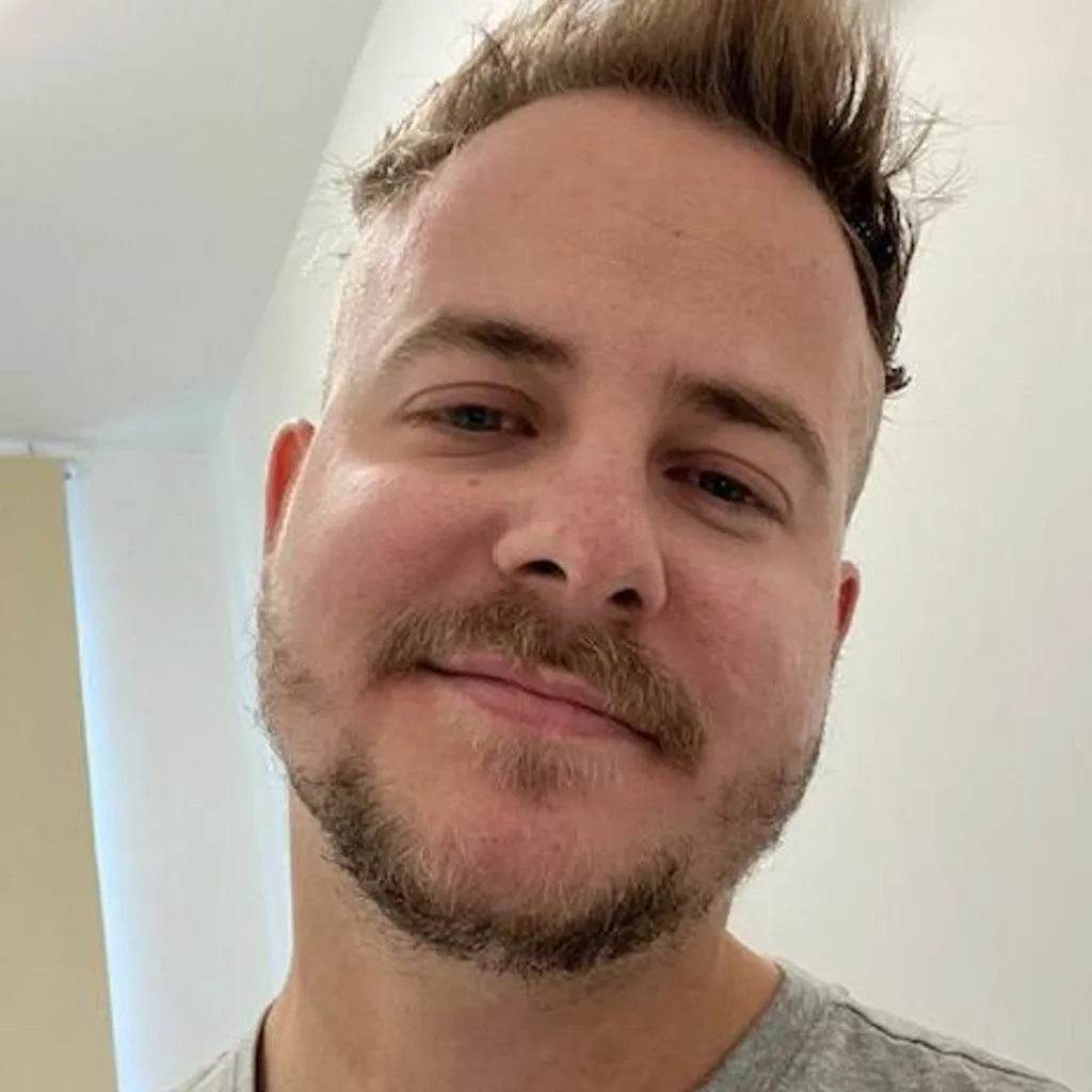 James wearing a light grey T‑shirt standing indoors in a bright room. Part of a doorway or wall panel is visible behind him, with a window and a ceiling light in the background.