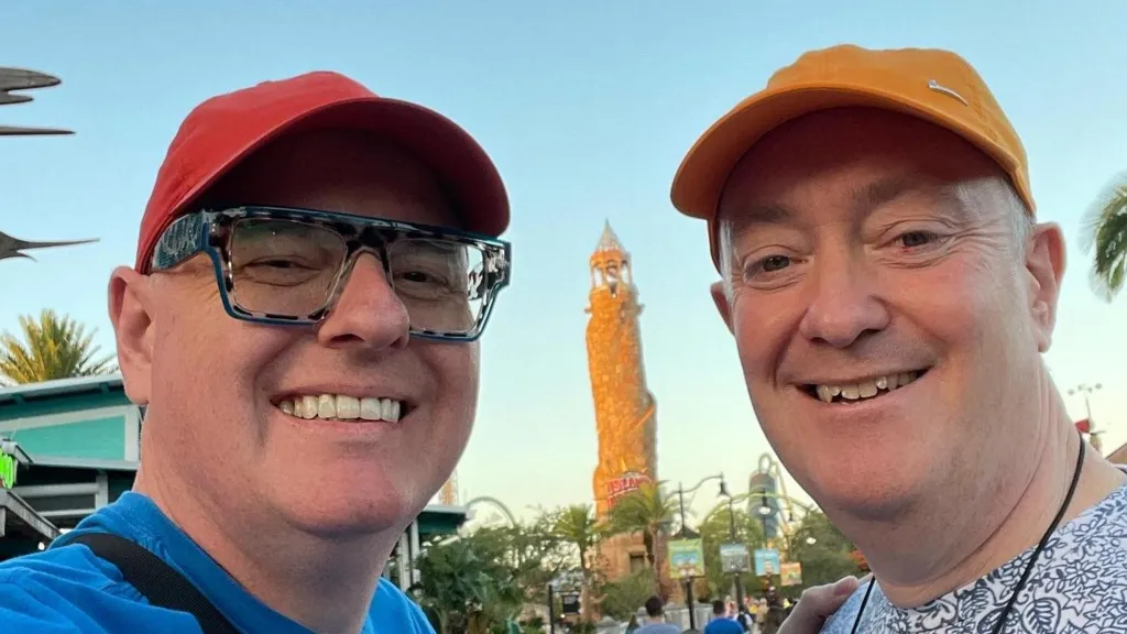 This is a selfie of Dave (on the left) and Cris (on the right) in a theme park. Both men are smiling. Dave wears a red baseball cap, black and white framed glasses and a blue t-shirt. Cris has a patterned blue and white t-shirt on and is wearing a mustard coloured baseball cap. 