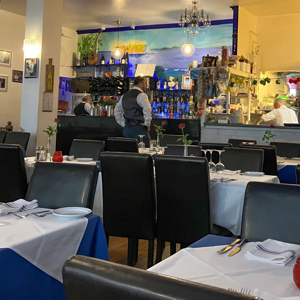 The interior of the Rozafa restaurant containing tables set with white cloths, cutlery, wine glasses, and red candle holders. Black chairs are arranged around the tables, and single red roses in vases are placed as centrepieces. Toward the back, staff members are working at a bar and open kitchen area with shelves of bottles and hanging utensils.