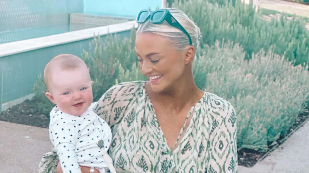 Kelly Medina Enos holding her daughter in her arms and smiling at her. She is in front of some plants