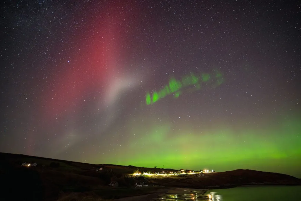 The Northern Lights in the north coast of Scotland. There are green and red lights across the sky with a village below. 