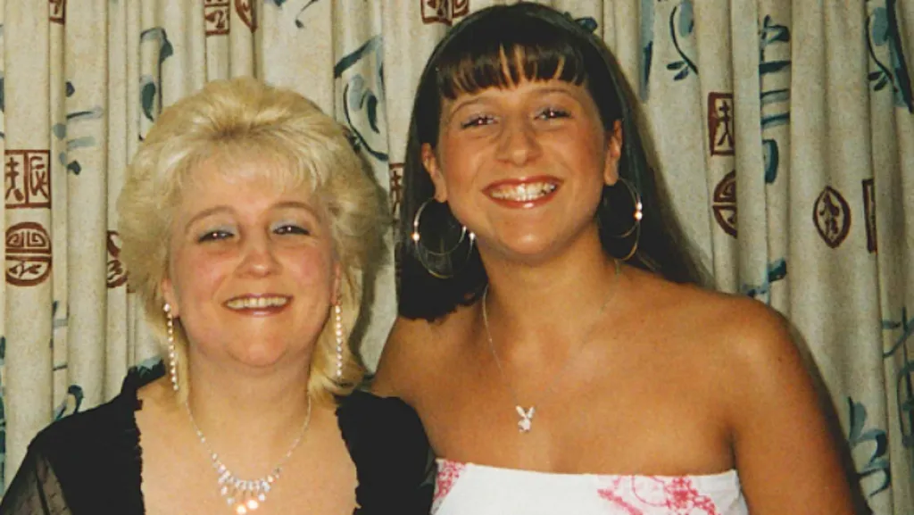 Pauline, in early middle age, and Jenna as a teenager. They are both dressed up. Pauline has layered, short blonde hair, is wearing makeup and diamante necklace and earrings. She has a black scoop neck top with ruffles at the edges and short, slightly see through lacy sleeves. Jenna has long dark hair and a fringe. She is tanned and wears a strapless white dress with red detailing either side. She has a number of very large hoop earrings and a silver chain with a pendant at the end. They are standing in front of curtains and both are smiling broadly.