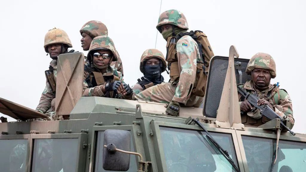 A group of six soldiers wearing camouflage uniforms and helmets riding on top of a large armored military vehicle. Some of them are holding rifles, and a few wear goggles or face coverings.