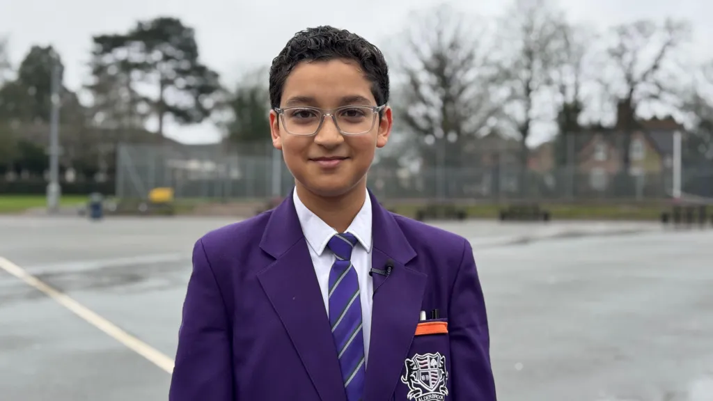 Youssef stands in the school grounds, wearing his uniform which is a purple blazer and tie and a white shirt. He has brown hair and brown eyes and wears glasses and is smiling at the camera.