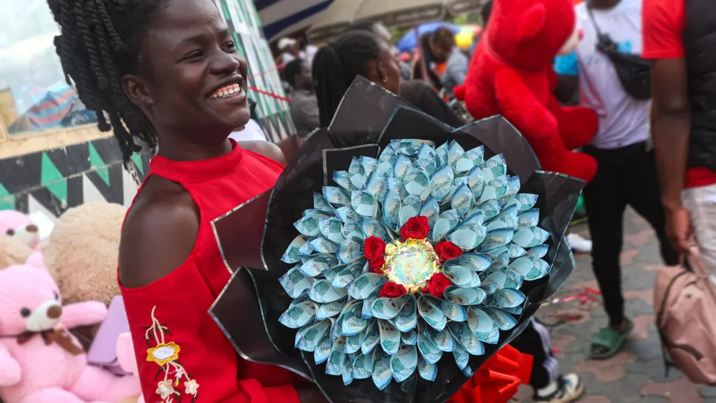 A woman in a market in Kenya holding a bouquet made out of blue coloured Kenyan shilling banknotes. Behind her is a stall selling teddy bears.
