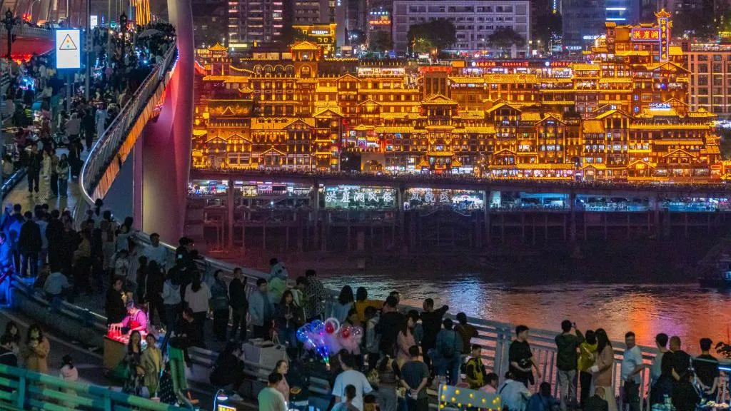 People crowding on a bridge overlooking a cluster of traditional-looking Chinese buildings, glowing in golden light.