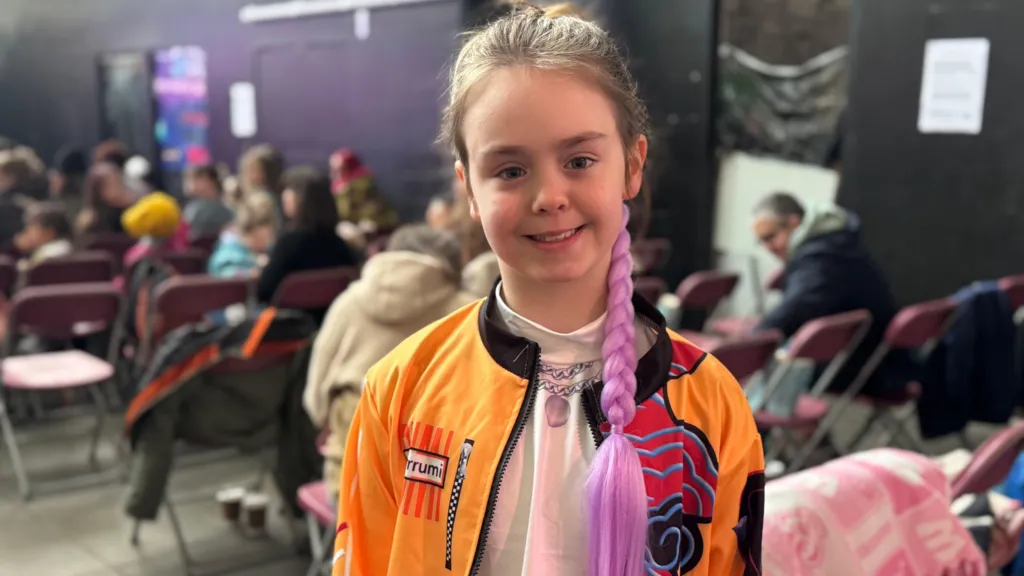 Phoebe is smiling at the camera. She is dressed as a Kpop character and is wearing an orange jacket. She has a purple ponytail.