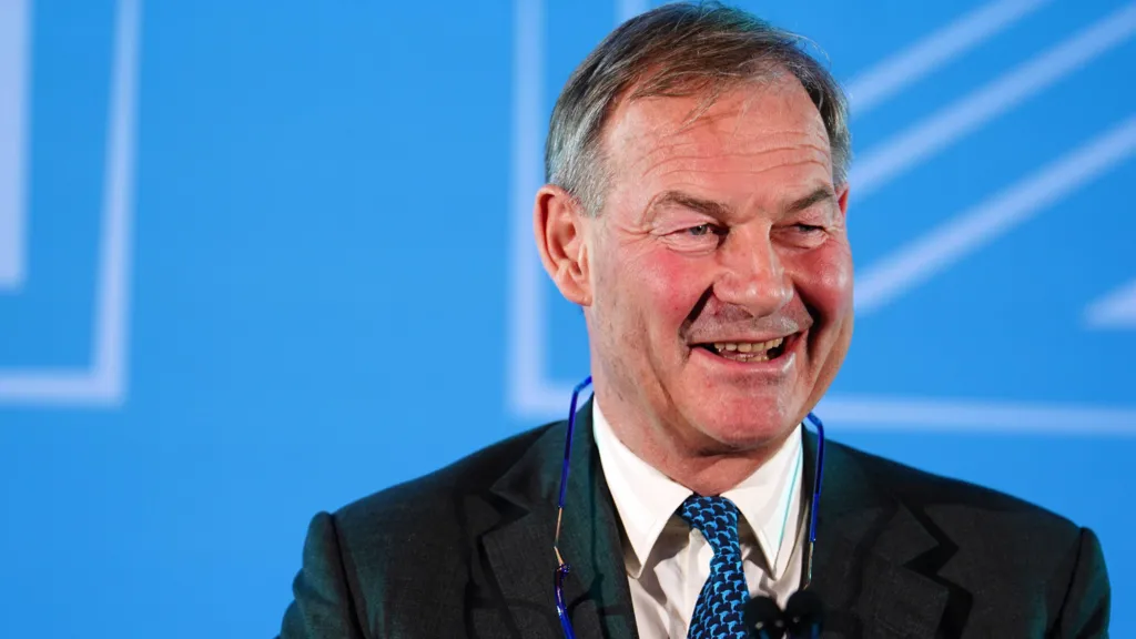 Rupert Lowe is wearing a suit blazer, a blue patterned tie and white shirt. He is smiling towards the right of the frame. There is a blue backing behind him.
