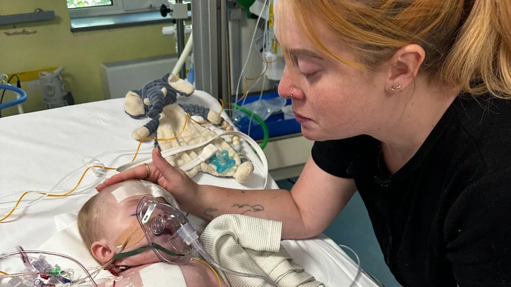 A baby and a mother looking over him. The mother has a black t-shirt on and ginger hair. The baby lies in the hospital bed and has tubes on his face and body.