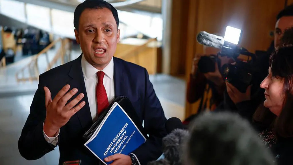 Anas Sarwar in blue suit, white shirt and red tie is holding a document bearing the words "Queen Elizabeth University Hospital" and gesticulating while talking to a crowd of journalists. They and their cameras are huddled around him. They are in the lobby of the Scottish Parliament building at Holyrood, which features light wood and grey concrete floors.