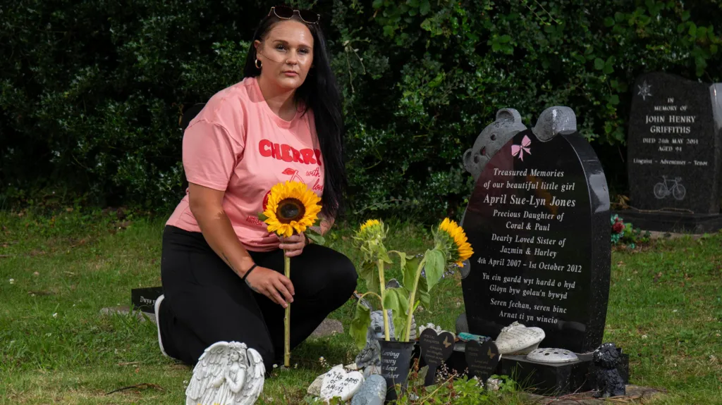 Photograph of Hazel Jones leaning in front of April's gravestone in Machynlleth. She holds a bright yellow sunflower in her hands and wears a pink t-shirt which reads "cherry". She has long black hair and blue eyes.