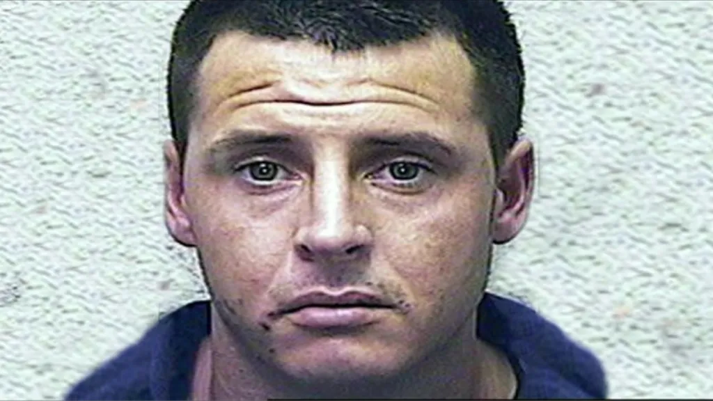 Police headshot of Jason Shaddick. He is a white man in his 20s with close cropped dark brown hair and green eyes. He is wearing a blue top. He is unsmiling and looking straight at the camera.
