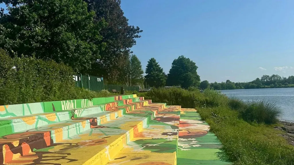 River Dee in sunshine. A pavement runs next to the riverbank and a bench, bridge and life ring can be seen.