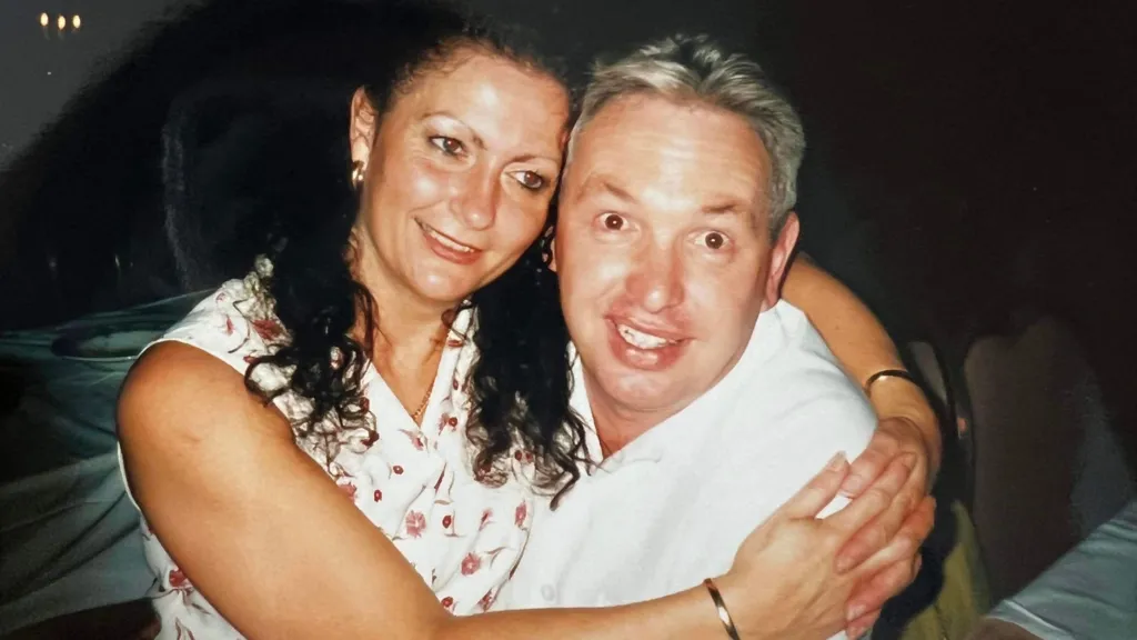 Joy, who has long curly hair and wears a sleeveless floral dress, in the arms of a man wearing a white shirt