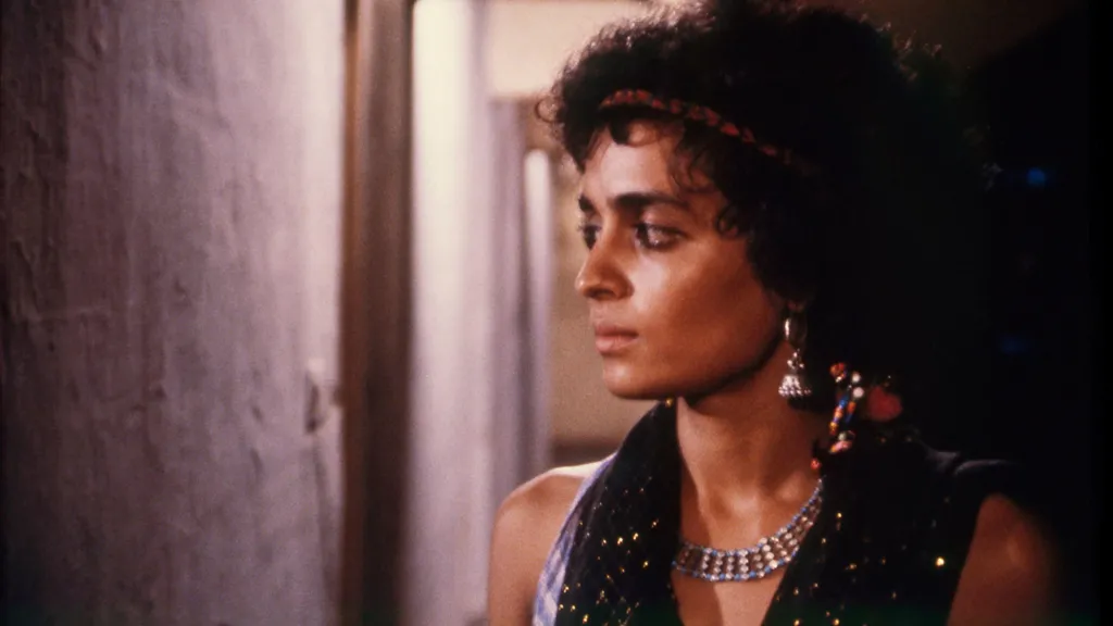 A still of Arundhati Roy, wearing silver danglers and a necklace and a black tank top, as she looks sideways 