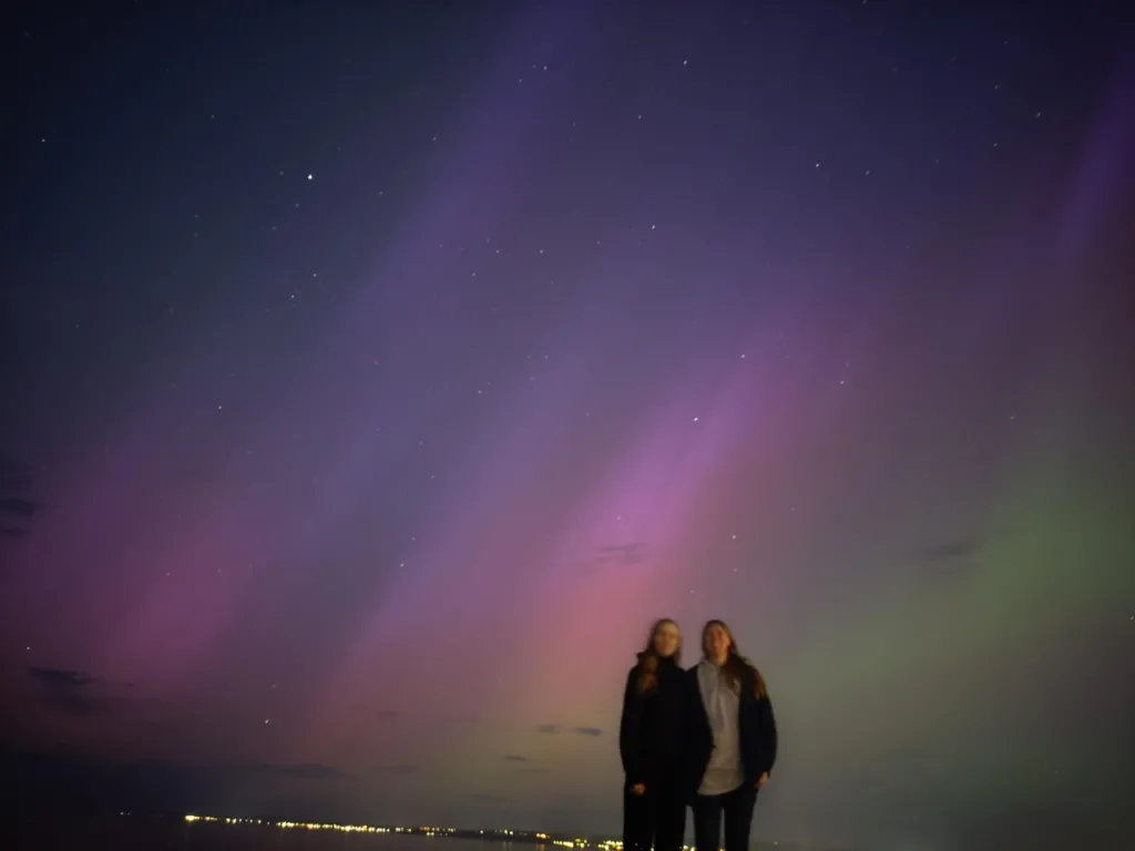 The Northern Lights from Portobello Beach in Scotland. There are green and purple stripes across the sky with Catherine Heymans and her teenage daughter in the foreground. 