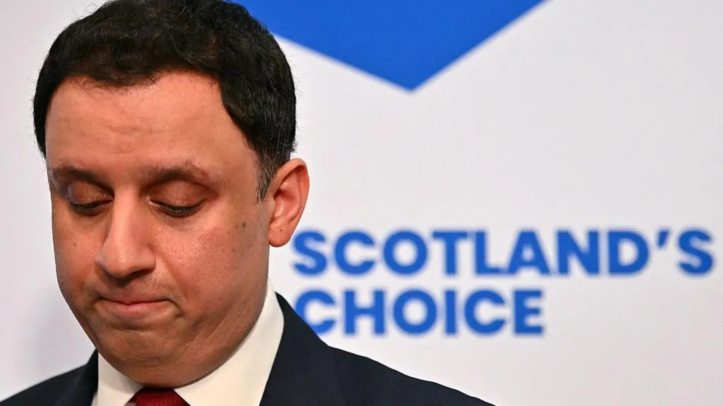 Anas Sarwar in dark suit and white shirt with red tie looks down as though in thought. A blue and white saltire backdrop with the words "Scotland's Choice" is directly behind him.