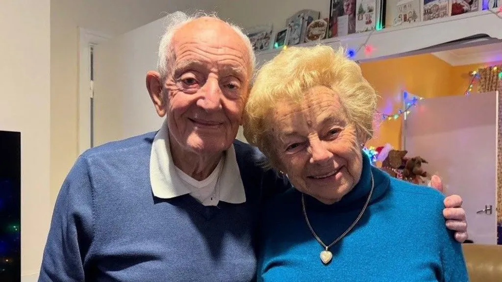 An older gentleman wearing a blue sweatshirt has his arm around his wife's shoulders, she is smiling in a light blue jumper wearing a gold locket around her neck
