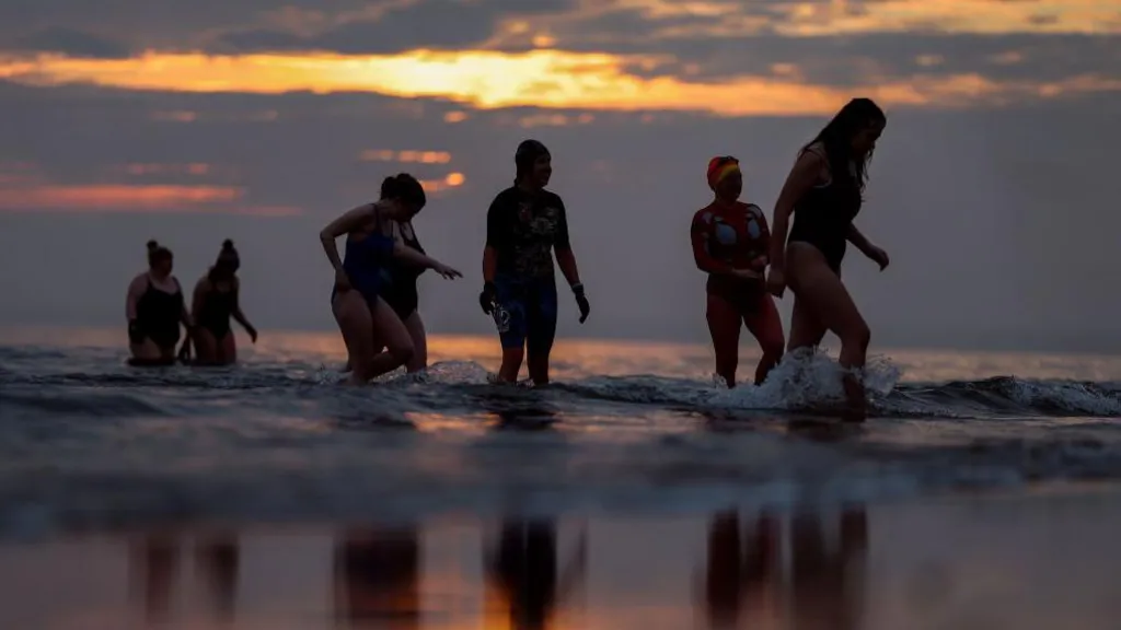 Seven women in swimming costumes wading pout of the sea at sunrise