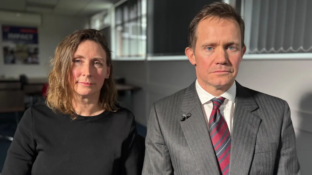 Eileen Teeton in a black top and shoulder length brown hair, sits next to her husband Mark, who has short hair and is dressed in a grey suit.