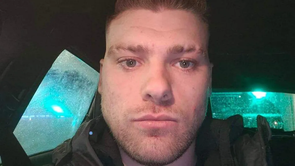 A close-up of Brandon Rainey, a man with short, fair hair and stubble, looking at the camera. He is sitting in a vehicle on a dark, wet night.
