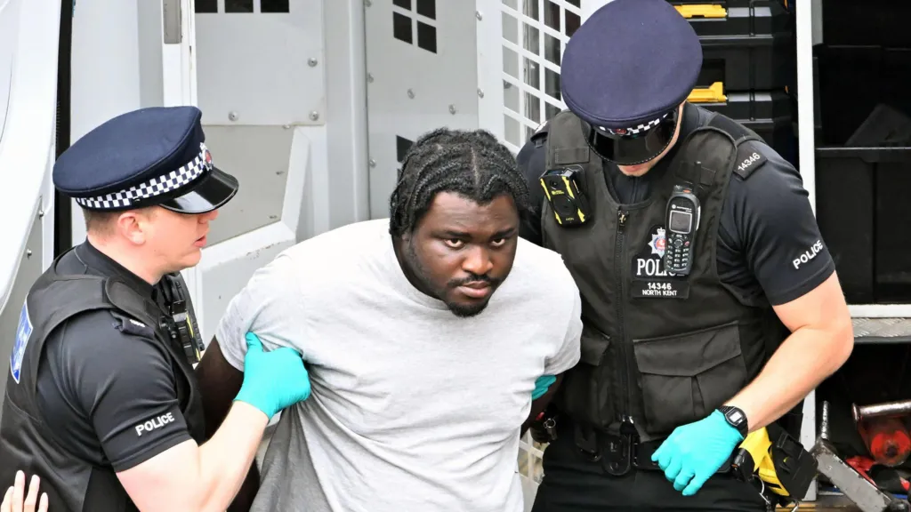 Anthony Esan is seen being held by two police officers, who are wearing uniforms and gloves. Esan is wearing a grey top and has his hands behind his back.