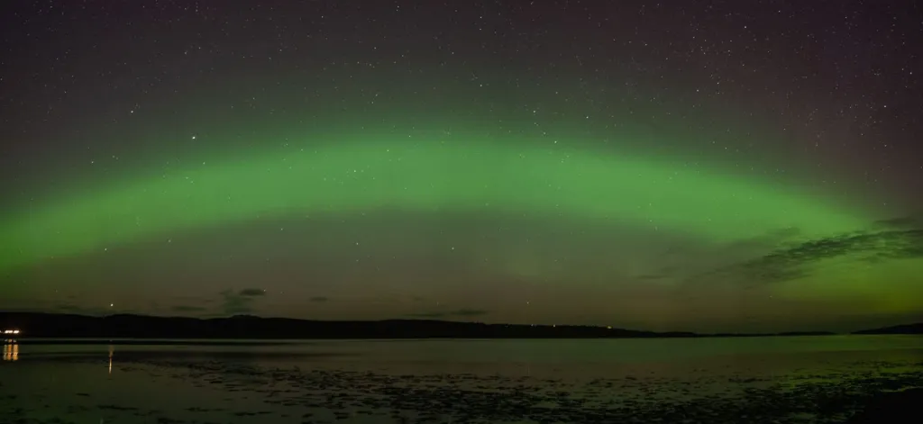 The Northern Lights in the north coast of Scotland. There is a green arc in the sky with the sea and mountains below. 