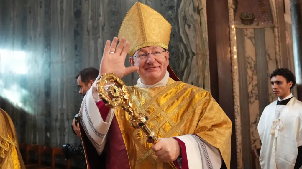 Bishop Richard Moth during his installation Mass as the 12th Archbishop of Westminster. He is smiling as he waves at onlookers. He is dressed in a gold robe and carries a gold crozier (pastoral staff)