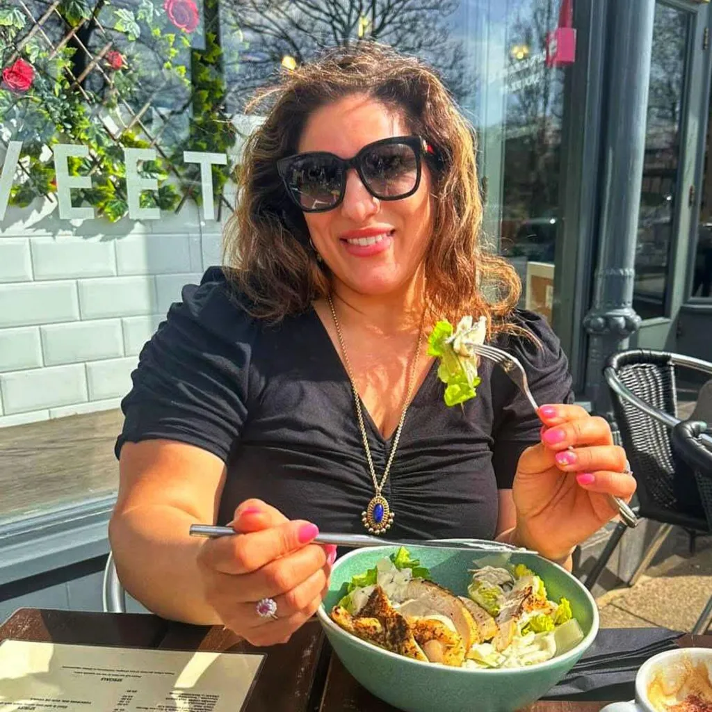 Vicki sitting at an outdoor table eating a bowl of salad. She is holding a fork with a bite of food and has a cup of coffee, cutlery, and a menu on the wooden table. The restaurant’s exterior has large windows and decorative flowers are visible behind them.