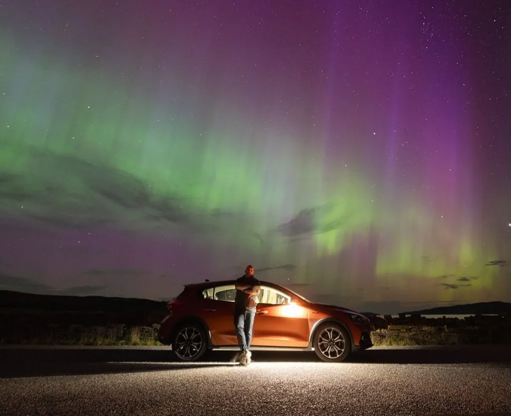 The Northern Lights in the north coast of Scotland. There are green and purple ripples across the sky. Gary Macleod stands in front of his car under the lights.