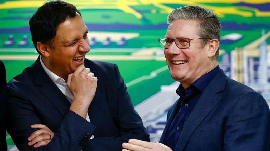 Anas Sarwar and Keir Starmer stand side-by-side, left and right of shot. The backdrop appears to be a very colourful technical graphic. Both men are in blue suits, Sarwar has a white shirt, Starmer has a dark blue one. They are not wearing ties and are laughing together.