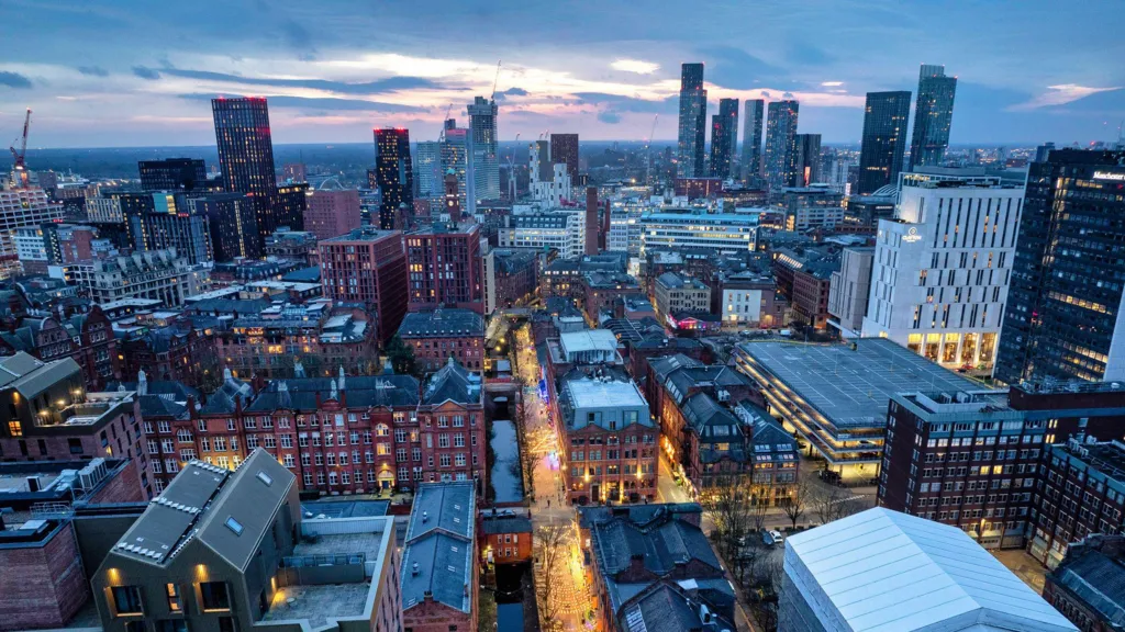 Manchester's skyline in the early evening with many modern skycrapers and cranes in the distance and older redbrick buildings in the foreground and at street level