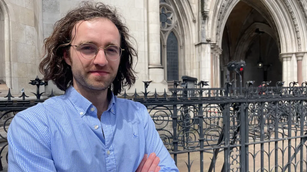 A man with long brown wavy hair and thin gold-rimmed glasses stands outside the gates at the Royal Courts of Justice in London. He is wearing a blue and white checked shirt and has his arms folded. He is looking directly into the camera.