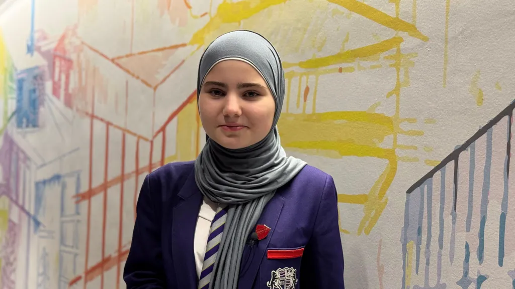 Year 11 student Jamila stands in front of a multi-coloured mural. She has a headscarf on and wears the school's purple blazer over a white shirt. She has brown eyes and is smiling.